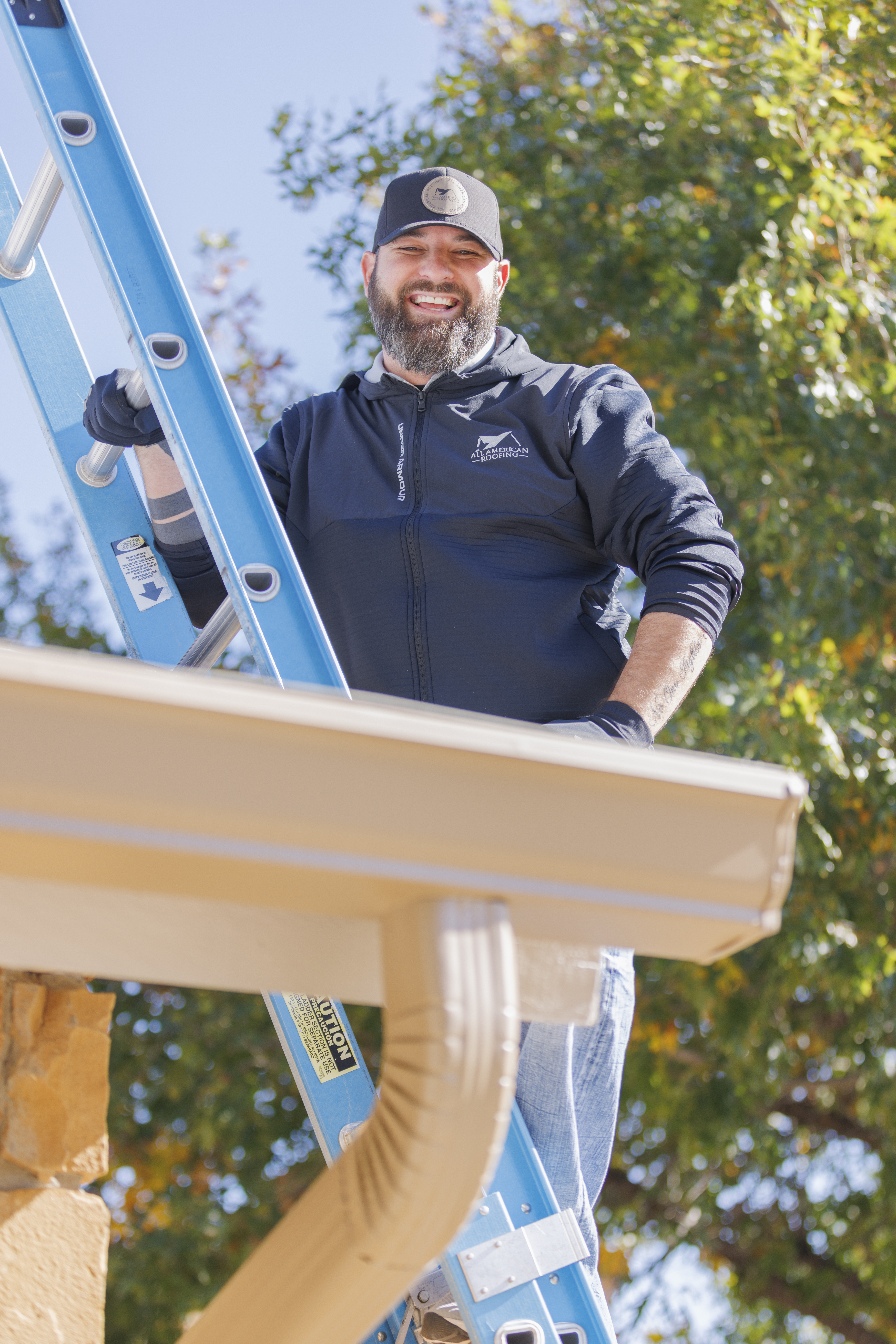 Smiling roofing professional standing on a blue ladder next to a house, wearing a black cap and jacket with the All American Roofing logo. The worker is positioned near beige rain gutters under a clear blue sky, with leafy green trees in the background.