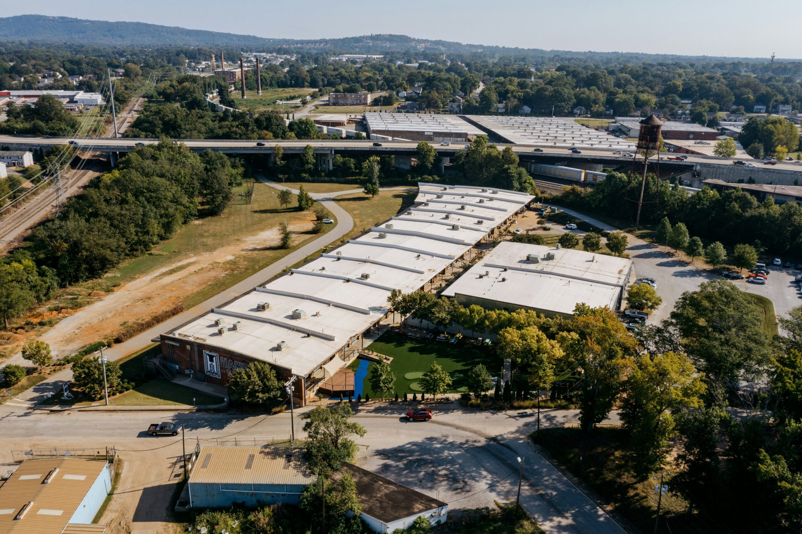 Aerial view of an industrial complex with flat-roofed buildings, surrounding trees, nearby railroad tracks, and a distant water tower.